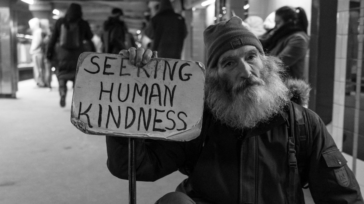 Homeless man holding a sign that reads "seeking human kindness"