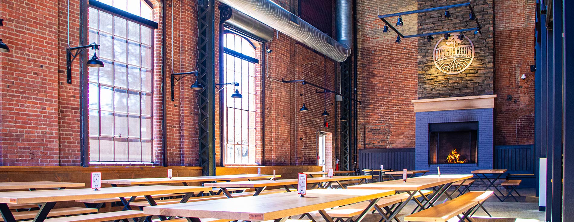 Large room with massive stone fireplace, brick walls, tall windows, and long wooden tables